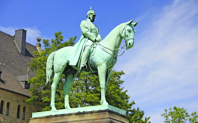 Reiterdenkmal aus Metall vor einem Gebäude mit blauem Himmel im Hintergrund.