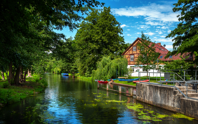 Haus am Fluss mit Booten und Bäumen an einem sonnigen Tag.