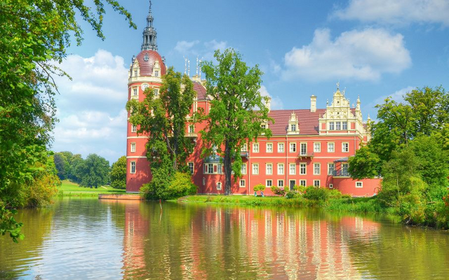 Rot-weißer Palast am Wasser mit Bäumen, blauem Himmel und Wolken im Hintergrund.