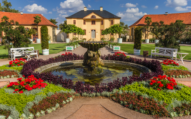 Historischer Garten mit Springbrunnen, umgeben von Blumen und Gebäuden im Barockstil.