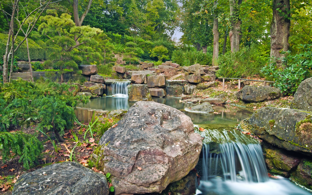 Wasserfall in einem grünen, felsigen Garten mit Baumkulisse.