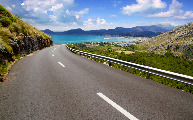 Straße in Landschaft mit Blick auf Küste und Berge bei klarem Himmel.