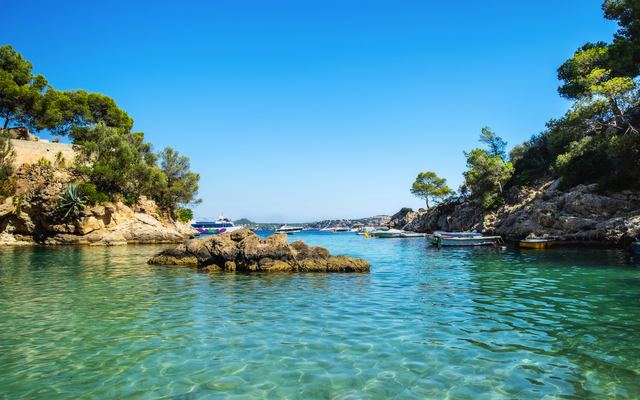 Klares blaues Wasser in einer Bucht mit Felsen und Pinienbäumen unter blauem Himmel.