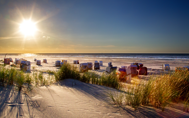 Sonnenuntergang an einem Sandstrand mit Strandkörben und Dünengras im Vordergrund.