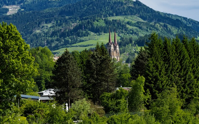 Kirche zwischen grünen Bäumen vor einer Bergkulisse.