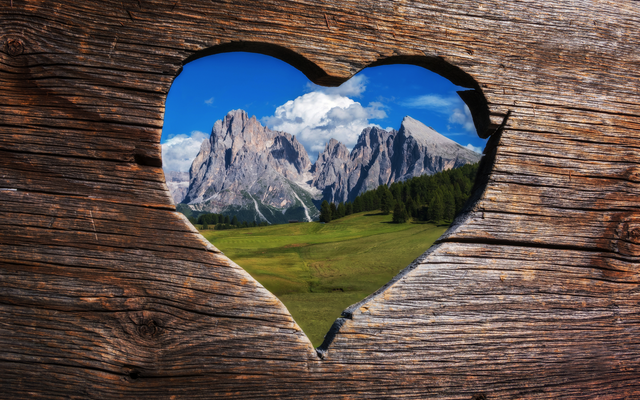 Herzförmiger Ausschnitt in Holz, zeigt Berge und Wiesen.