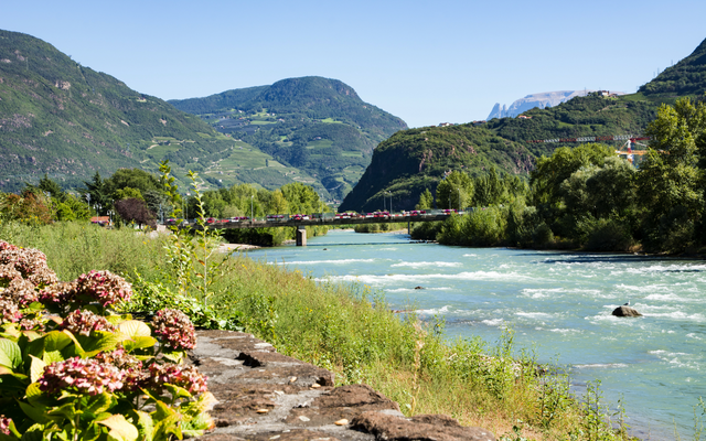 Fluss in einer bergigen Landschaft mit Brücke und grünem Ufer im Vordergrund