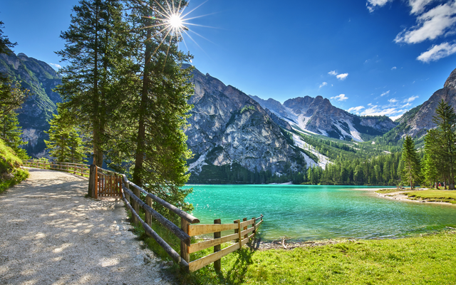 Bergsee in den Alpen mit Waldweg und klar blauem Himmel.