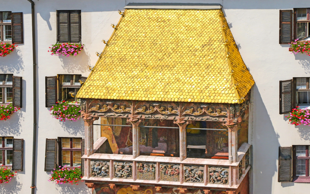 Balkon mit goldenem Dach und bunten Blumen an Fenstern einer weißen Fassade.