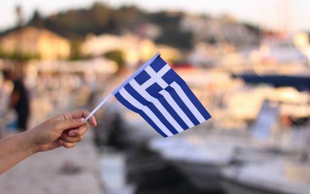Hand, die eine griechische Flagge vor einem Hafen hält.