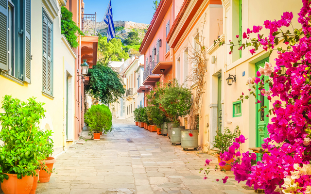 Bunte Gasse mit Blumen und griechischer Flagge im Hintergrund.