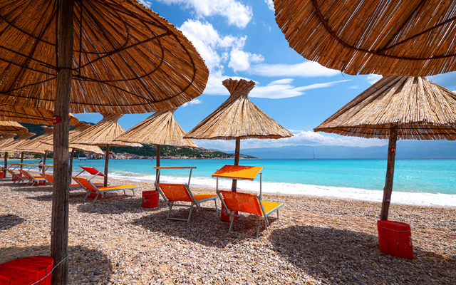 Strand mit Sonnenliegen und Strohdächern unter blauem Himmel.