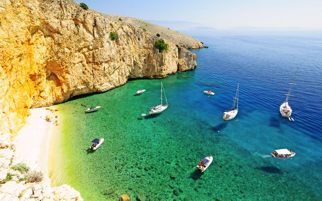 Segelboote auf klarem, türkisfarbenem Wasser entlang einer felsigen Küste mit goldenem Sandstrand.