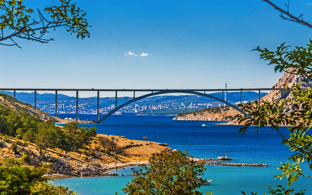 Brücke über türkisblaues Meer mit Stadt im Hintergrund und Bäumen im Vordergrund.