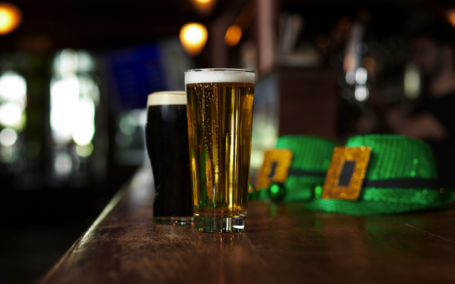 Zwei Gläser Bier auf einem Holztresen mit grünen Hüten im Hintergrund.