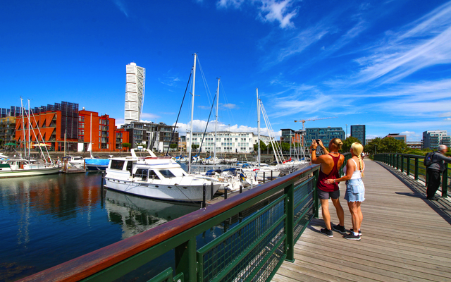 Personen auf einer Promenade mit Blick auf Yachthafen und moderne Gebäude im Hintergrund.