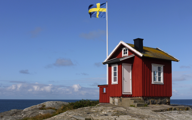 Rotes Häuschen auf Felsen, schwedische Flagge weht im Wind, Meer im Hintergrund.