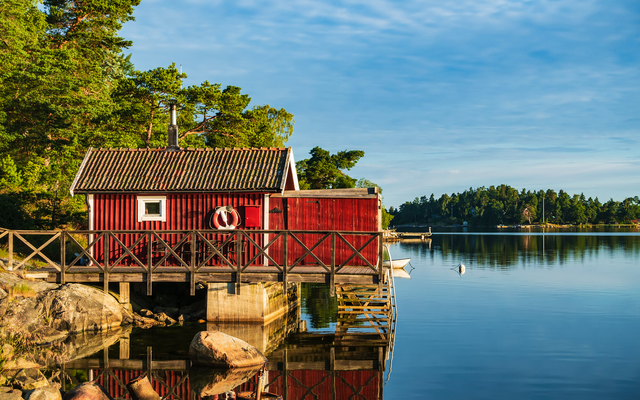 Rotes Holzhaus am Ufer eines Sees mit Bäumen im Hintergrund.