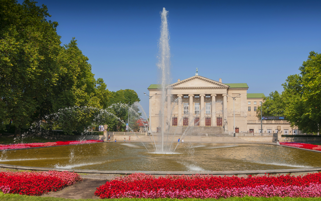 Springbrunnen vor einem großen Gebäude mit Säulen und blühender Landschaft.