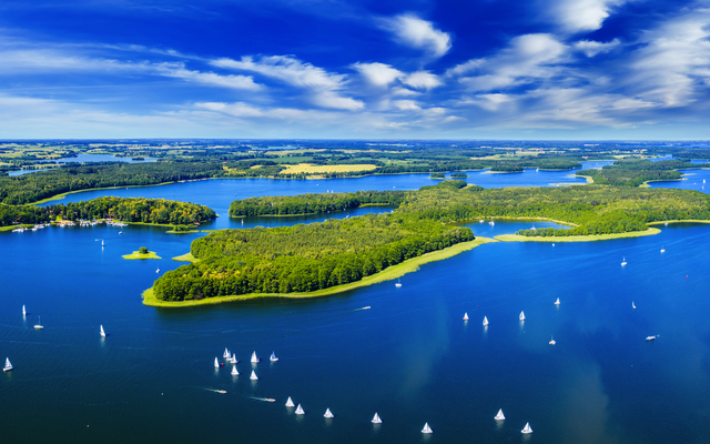 Luftaufnahme eines Sees mit Inseln und zahlreichen Segelbooten unter blauem Himmel.