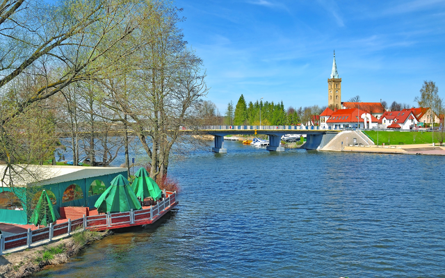 Flusslandschaft mit Brücke und Kirchturm, Bäume und Gebäude am Ufer.