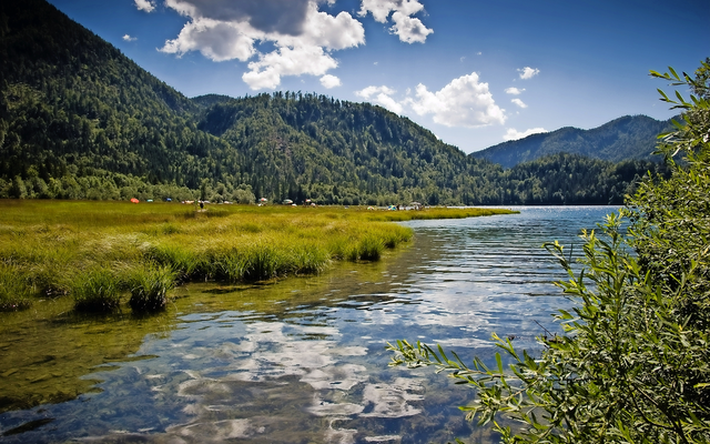 Bergsee in bewaldeter Landschaft unter blauem Himmel mit weißen Wolken.