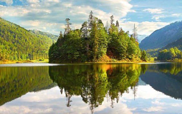 See mit bewaldeter Insel und Bergen, Spiegelung im Wasser, blauer Himmel mit Wolken.