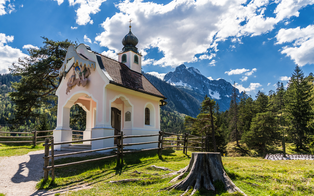 Kapelle in alpiner Landschaft, umgeben von Wiesen und Bergen unter blauem Himmel.