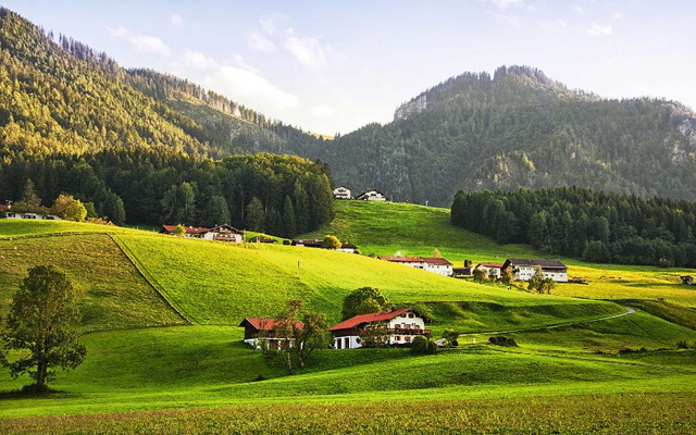Berglandschaft mit grünen Wiesen und vereinzelten Häusern vor bewaldeten Hügeln.