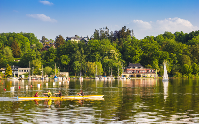 Vierer-Ruderboot auf einem See vor einer grünen Uferlandschaft mit Segelboot.