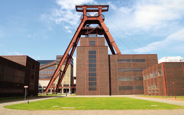 Industriegebäude mit rotem Förderturm im Vordergrund auf grünem Gelände.