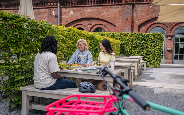 Gruppe von Menschen sitzt an einem Tisch im Freien vor Backsteingebäude.