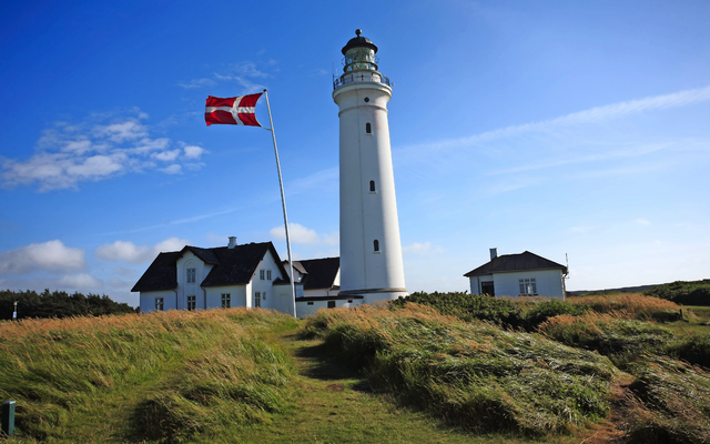Leuchtturm mit dänischer Flagge vor blauem Himmel und weißen Gebäuden im Hintergrund.