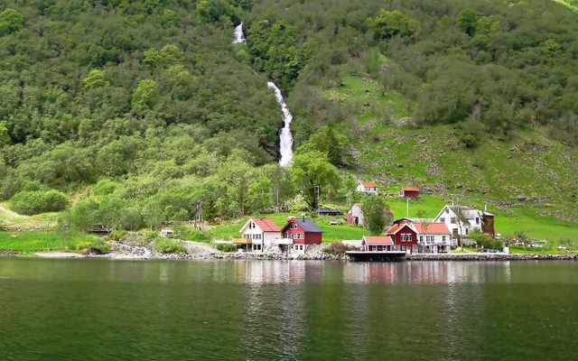 Häuser am Ufer eines Fjords mit Wasserfall im Hintergrund.