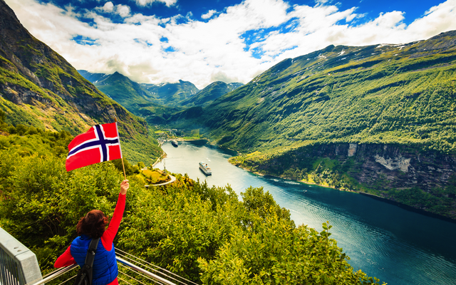 Person schwenkt norwegische Flagge vor Fjordlandschaft.
