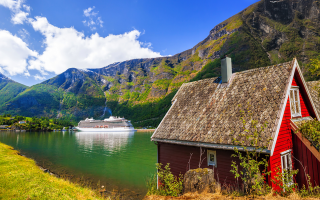 Rotes Haus am Fjord mit Kreuzfahrtschiff und Bergen im Hintergrund bei Sonnenschein.