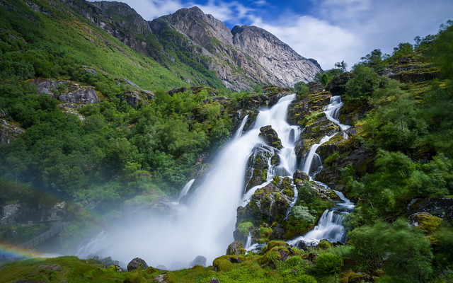Wasserfall in bewaldeter Gebirgslandschaft mit Regenbogen am Fuße.