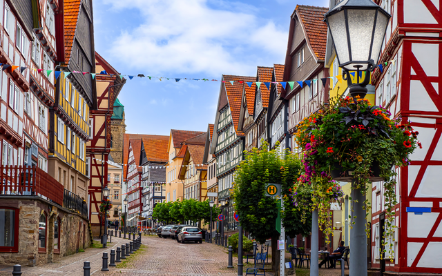 Gepflasterte Straße mit Fachwerkhäusern und Blumen an einer Laterne.
