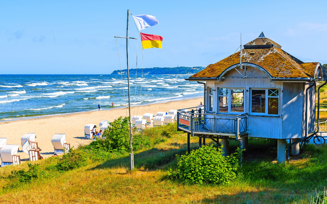 Strand mit Strandkörben, Haus am Meer und wehenden Flaggen.