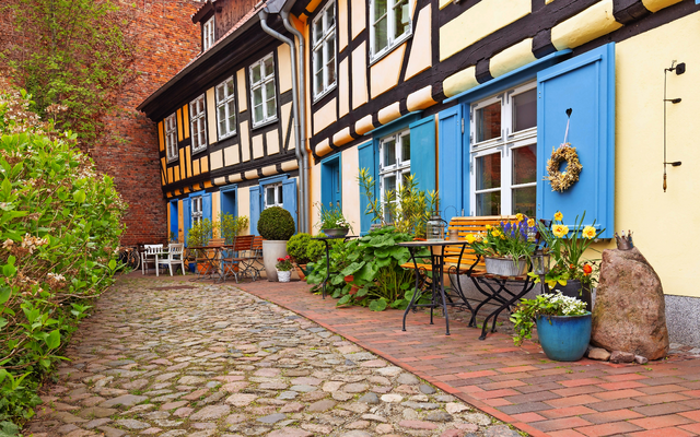 Fachwerkhaus mit blauen Fensterläden, Blumendeko und Kopfsteinpflaster in schmalem Innenhof.