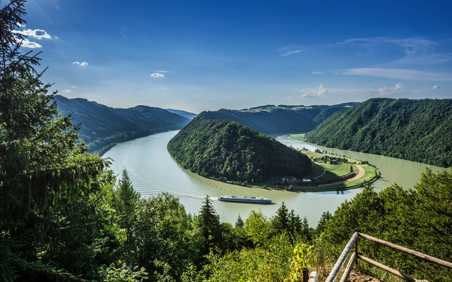 Flusslandschaft mit Wald und Schiff unter blauem Himmel.