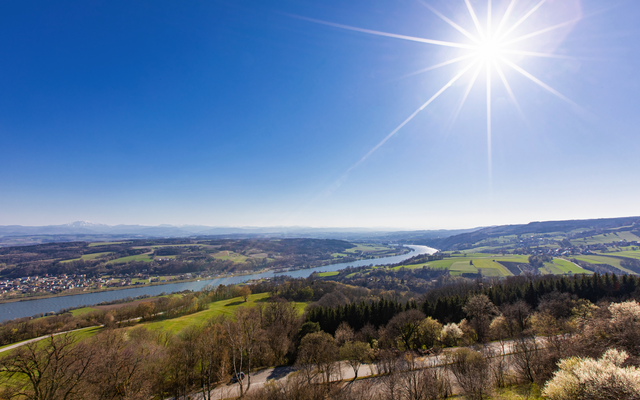 Landschaft mit Fluss, Feldern, Wäldern und strahlender Sonne am blauen Himmel.