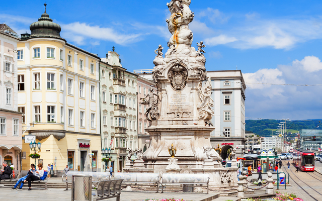 Barocke Dreifaltigkeitssäule auf Stadtplatz mit Blumendekorationen und umliegenden Gebäuden.
