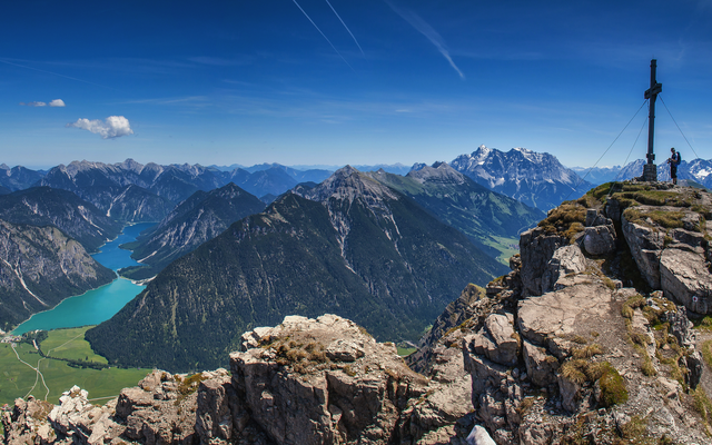 Bergpanorama mit Gipfelkreuz und Tal mit türkisfarbenem See unter blauem Himmel.