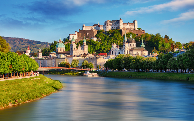 Festung Hohensalzburg über der Stadt Salzburg mit Fluss im Vordergrund bei Sonnenuntergang.