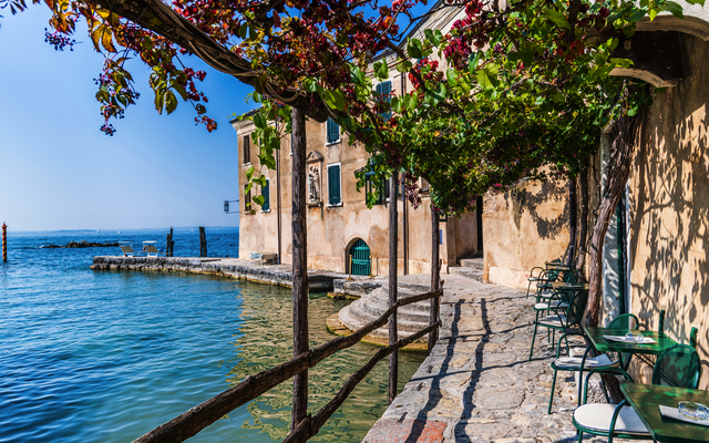 Bewachsener Weg mit Blick aufs Wasser und Gebäude im mediterranen Stil.