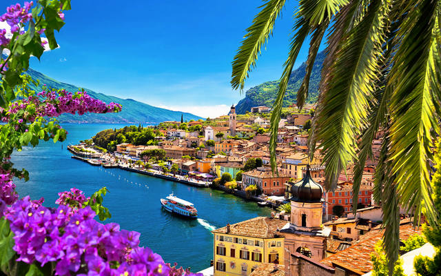 Mediterrane Küstenstadt mit Palmen und blühenden Blumen, im Hintergrund ein See.