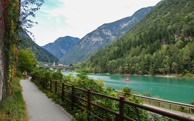 Radweg entlang eines Sees mit Bergen und Wald im Hintergrund.