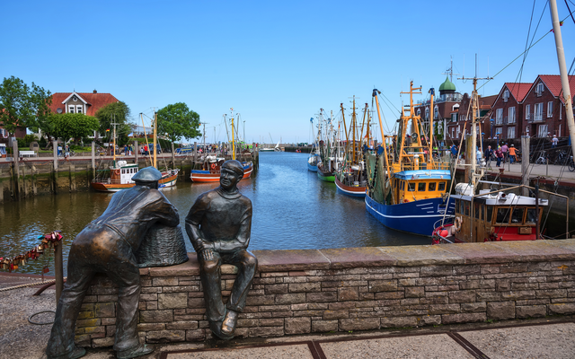 Hafen mit Fischkuttern, Backsteinhäusern und Bronze-Skulpturen im Vordergrund.