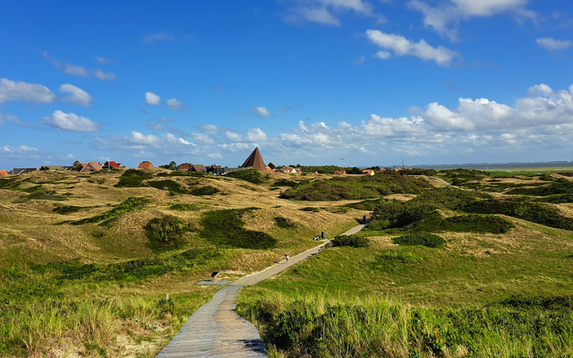 Holzweg durch grasbewachsene Dünen unter blauem Himmel mit Wolken.